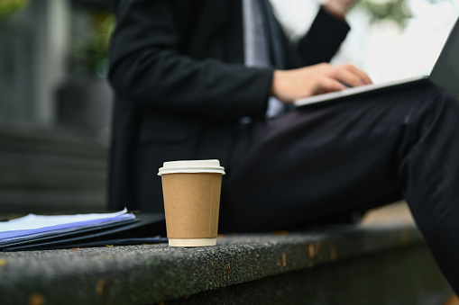 A disposable cup of coffee on stairs outside office building with businessman using laptop in background.