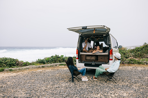 A couple is laying in the bed of the campervan while having a conversation with their two friends who are sitting outside