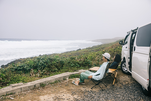 Japanese couple sitting and smiling next to their campervan while enjoying their view of the ocean