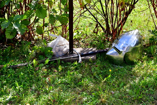 A close up on an old medieval helmet made out of steel or brass located next to a one handed sword and a bag of potatoes stored in a foil bag seen in the garden on a sunny summer day in Poland