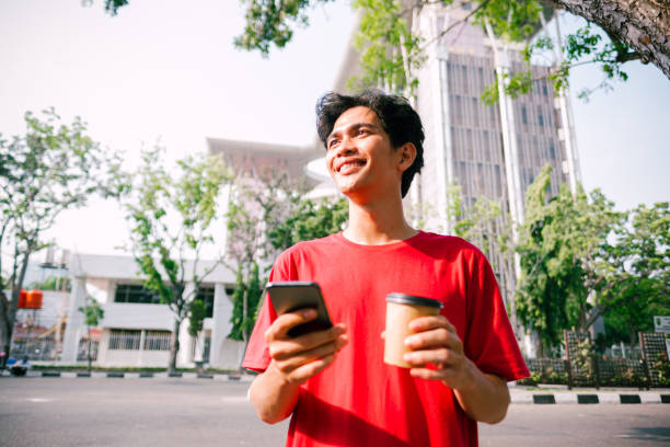 Young Adult Asian Man Smiling While Holding Smartphone At Sidewalk.