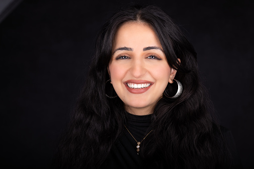 Middle eastern young woman in a studio headshot over black background. She is happilly smiling to the camera