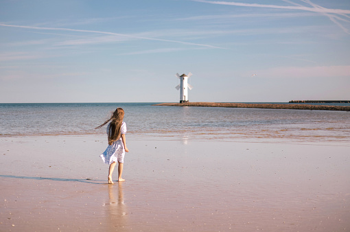 Back view of adorable little girl with long hair in white dress walking on tropical beach vacation