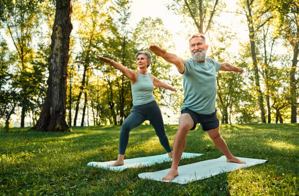 Active retirement life. Determined family couple standing on rubber mats in national park and performing warrior asana pose. Aged man and woman showing strength and flexibility when practicing yoga. yoga stock pictures, royalty-free photos & images