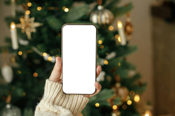 istock Christmas phone mock up. Hand holding smartphone with empty screen against stylish festive christmas tree with golden lights. Space for text. Christmas advertising, smartphone app template 1716541475