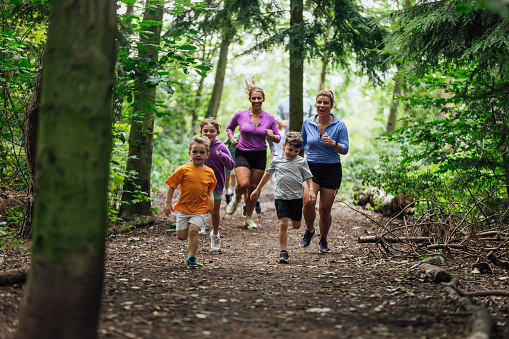 A wide shot of a group of adults and children wearing sports clothing. They are taking part in a fun run with other children and adults at Cramlington Nature Reserve in the North East of England. Two women and three children run down a footpath in a woodland.