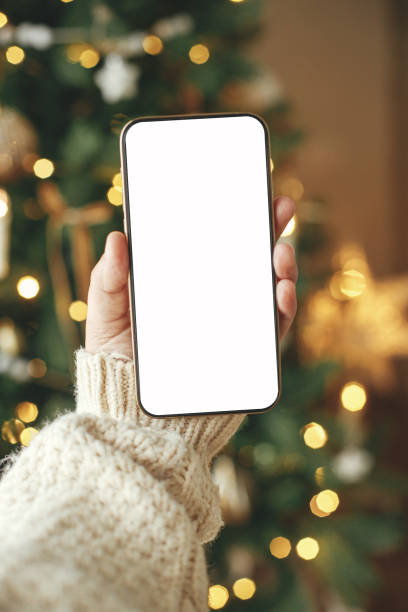 Hand holding smartphone with empty screen against stylish festive christmas tree with golden lights.Christmas phone mock up. Space for text. Christmas advertising, smartphone app template