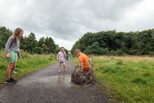 A wide shot of four children wearing sports clothing on an overcast day in a nature reserve in the North East of England. They are jumping and splashing in a puddle on a footpath.