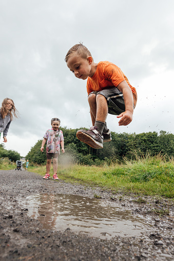 A medium, low-angle shot of three children wearing sports clothing on an overcast day in a nature reserve in the North East of England. They are jumping and splashing in a puddle on a footpath.