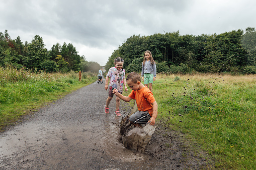 A wide shot of three children wearing sports clothing on an overcast day in a nature reserve in the North East of England. They are jumping and splashing in a puddle on a footpath.