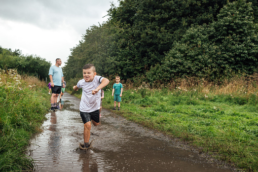 A wide shot of three children and a mid-adult man wearing sports clothing on an overcast day in a nature reserve in the North East of England. They are jumping and splashing in a puddle on a footpath.