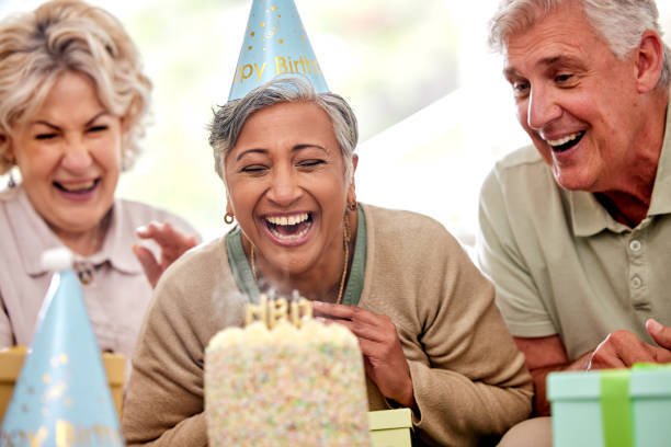 老人ホームで年配の女性、友人、誕生日パーティー、ケーキでろうそくを吹き、願い事とお祝いをする。高齢者グループ、人々、そして一緒に幸せに、イベントと引退の多様性で笑う - senior adult nursing home eating home interior ストックフォトと画像