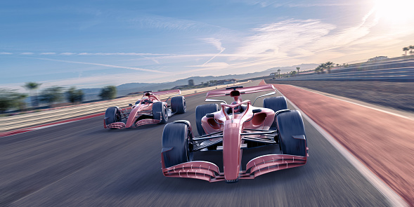 Two generic red racing cars with same livery driving at high speed along a straight section of racetrack under bright blue dawn sky with some white clouds.