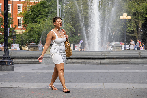 Washington Square Park, Manhattan, New York, USA - August 16th 2023:   Young woman in a summer dress passing the fountain in the center of the park