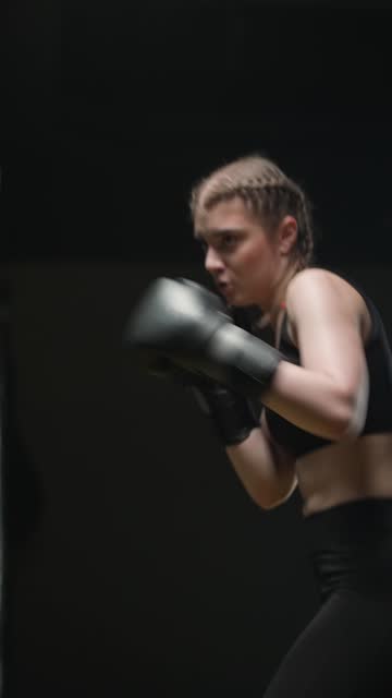 Woman fighter trains his punches and defense in the boxing gym, the girl strikes fast, beats a punching bag, vertical video.