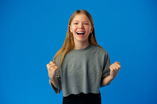 Portrait of a joyful happy teenage girl celebrating success on blue background in studio