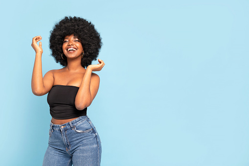 Portrait of cheerful young African American female model in black top and jeans, with afro hairstyle, smiling to the camera. Real people emotions. A lot of copy space. Blue pastel studio background.
