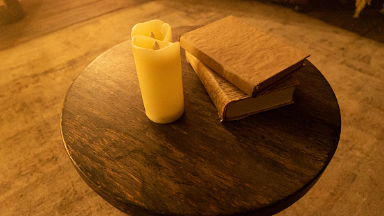 Flameless candles with vintage books on small table in room. Artificial candles and old foliants on round wooden desk in home interior. Simple details creating comfortable atmosphere indoors