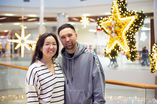 young Asian Kazakh male and female couple in shopping mall on Christmas or New Year holidays. happy family or man and woman in love against huge decor stars in public place. Valentine's Day, newlyweds