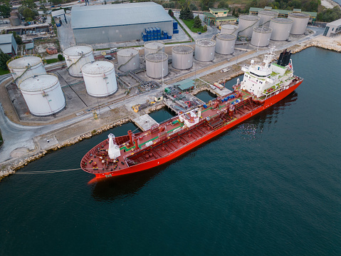 Tanker ship refueling at an oil terminal with storage silo's in the port, aerial view from flying drone