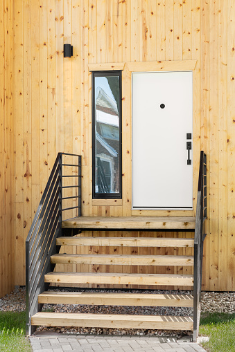 The front door of a home with a natural wooden siding and wrought iron railings and wood steps leading to a white front door.