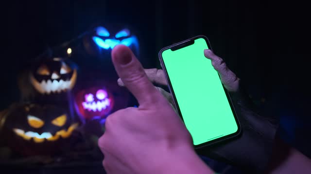 A girl in a witch costume shows like and holds a phone with a green screen on a Halloween background