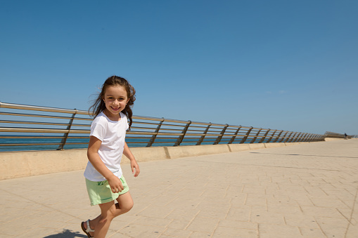 Beautiful little child girl in sporty clothes, smiles looking at camera while walking and playing in the Ocean promenade on a sunny warm summer day. Childhood. People. Leisure activity. Lifestyle