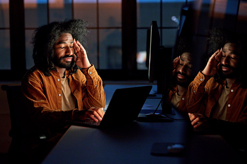 Waist-up view of bearded multiracial man in early 30s sitting at desk with eyes closed, face reflected and illuminated by computer screens.