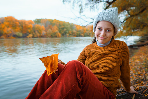 a young teenage girl posing in an autumn forest, sitting on a tree by the river bank, beautiful nature and bright yellow leaves