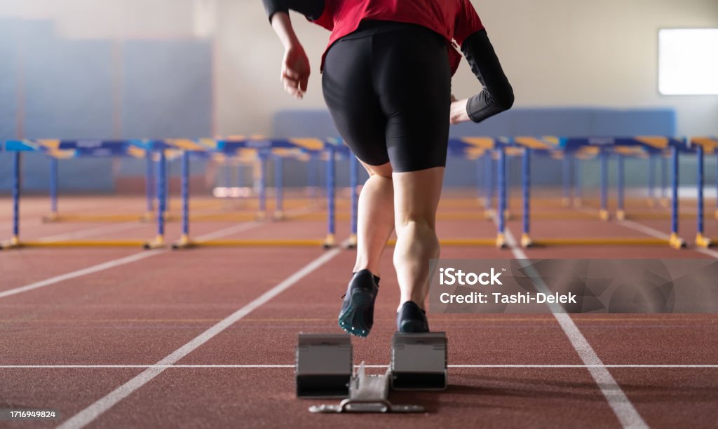 Female Runner Training And Jumping Over Hurdles Young female runner training and jumping over hurdles at indoors stadium running track. Sport and competition concept. Preparation Stock Photo