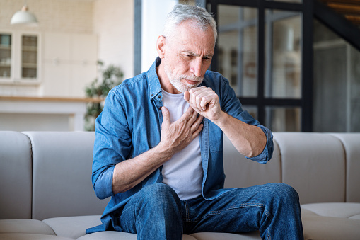 Mature man suffering from dry cough and chest pain while sitting alone on sofa in living room. Male feeling sick, has fever and respiratory infection symptoms. Healthcare concept