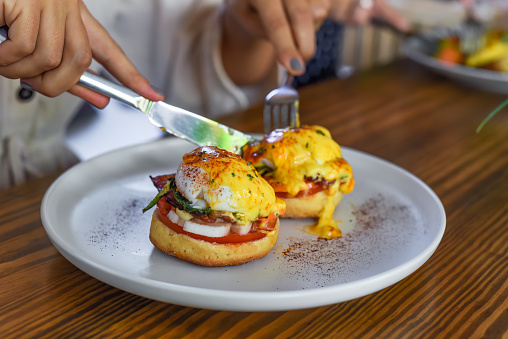 Hands with knife and fork cutting english muffin with poached eggs, bacon and Hollandaise sauce with vegetables