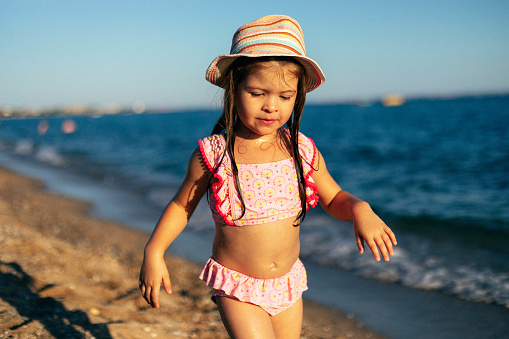 The three-year-old girl is walking by the sea. The child is wearing the swimsuit and a hat. The little girl is enjoying the beautiful evening on the beach.