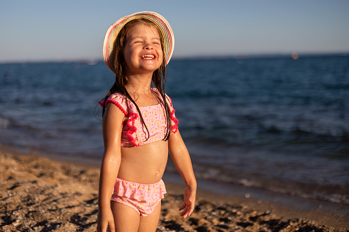 A child stands cheerfully on the seashore. The three-year-old girl wears a swimsuit and a summer hat and enjoys the beautiful sea and sunset.