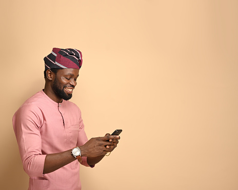 Title: Portrait of a handsome delightful African man wearing PNG of an African man in peach native, reading viral messages from his mobile