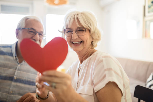 Tender Moments Smiling senior woman receives present box and kissing her husband at home senior birthday card stock pictures, royalty-free photos & images