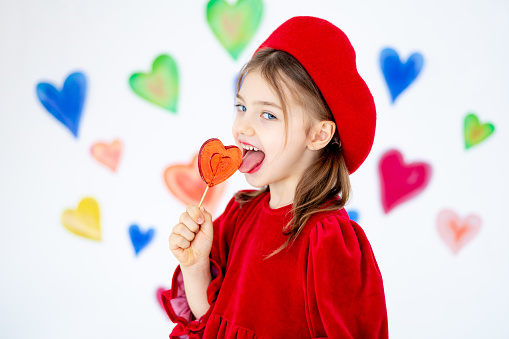 valentine's day, a small smiling child girl in a red beret happy holds or licks a large lollipop in the form of a heart and rejoices against the background of colorful hearts.