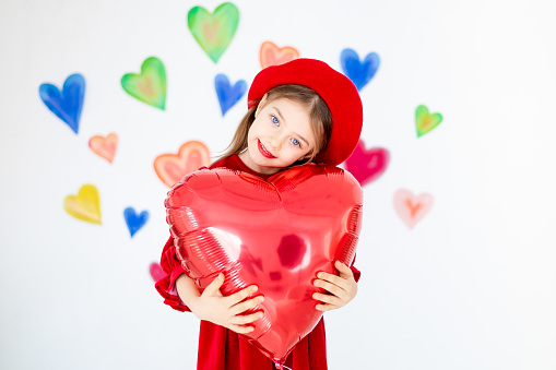 valentine's day, a small smiling child, a girl in a red beret, happy, holds a large red balloon in the form of a heart in her hands and rejoices against the background of colorful hearts.