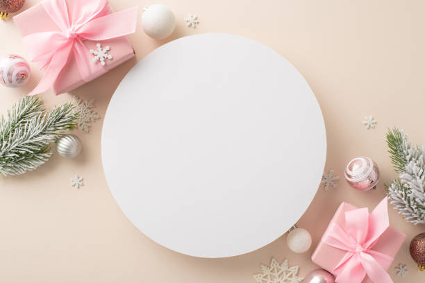 Frosty Holiday Composition: top view of pink gift parcels, delightful tree baubles, snowflakes covered in frost, pine boughs on beige backdrop. Customize empty circle with New Year's wishes or promos