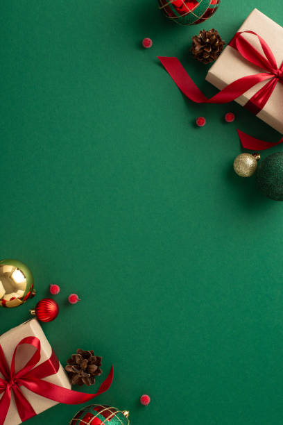istock Seasonal Splendor: vertical top view of kraft gift boxes with red ribbon bows, surrounded by chic tree toys, shimmering golden baubles, pine cone, mistletoe berries on green backdrop with ad space 1717118980