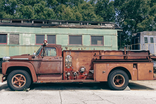 A rusty orange, 1950's fire truck is displayed in front of old train passenger cars. Abandoned collection at railroad museum in Lake Wales Florida