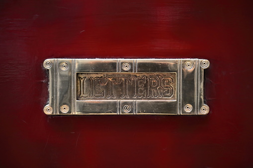 Brushed, brilliant metal flap cover of a mail slot cut through the lower half of a deep red-painted front door featuring the word ''letters'' chiseled on the metal lid. Melbourne-VIC-Australia.