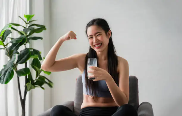 Portrait of happy Asian woman in sportswear sitting smiling and holding milkshake protein glass with pose showing her muscles on chair in the fitness room at home. Healthcare and weight loss concept.