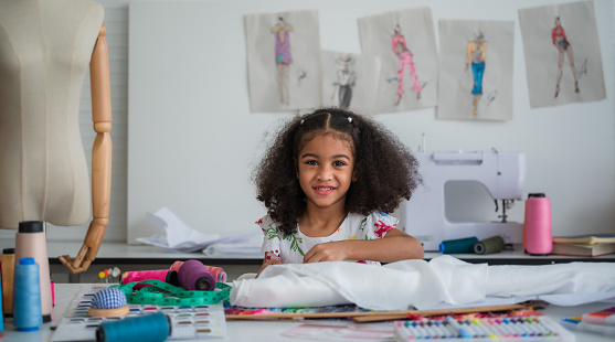 Portrait of a happy cute African little girl dressmaker with an afro hairstyle sitting sewing by handicraft and looking at camera in the fashion studio. Cheerful small child girl seamstress tailor.