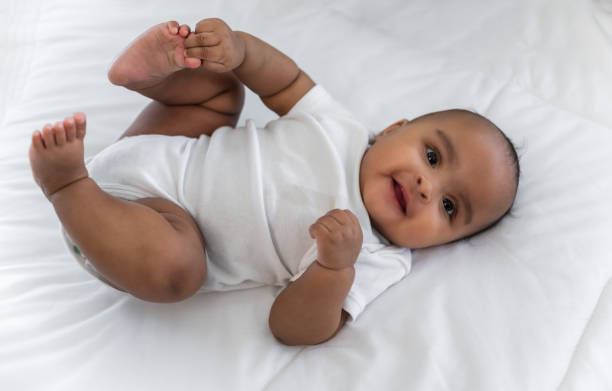 幸せなかわいい小さなアフリカの新生児のトップビューが笑顔で横になり、自宅の寝室の白いベッドでカメラを見る。 - newborn baby african ethnicity little girls ストックフォトと画像