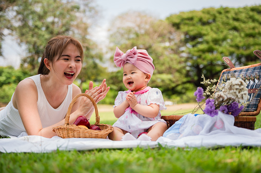 Portrait of happy Asian mother and her cute little daughter good mood smiling and playing having fun together while picnic in a public park. Cheerful adorable baby girl and her mom relaxing in garden.