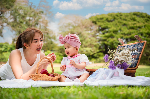 Portrait of happy Asian mother and her cute little daughter good mood smiling and playing having fun together while picnic in a public park. Cheerful adorable baby girl and her mom relaxing in garden.