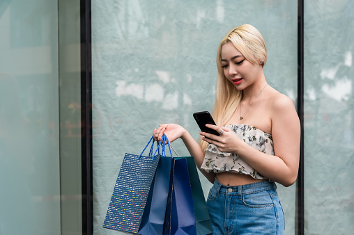 Portrait of happy beautiful Asian woman in clothes fashion with blonde hair standing holding shopping bags and using a smartphone at a shopping mall in city. Confident young female at a shopping mall.