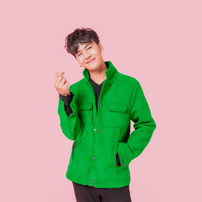 Portrait of happy Asian handsome man in fashionable green jacket clothing standing smiling and using fingers to making mini heart sign isolated on pink background. Young male fashion model lifestyle.