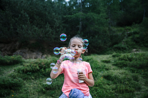 Little toddler girl blowing soap bubbles standing in beautiful green nature.
